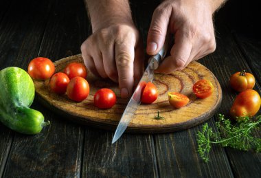Öğle yemeği için sebze yemeği hazırlamadan önce kesme tahtasındaki kiraz domatesi kesmek için bıçak kullanan bir adamın ellerini yakın plan çek..