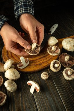 Bir adam lezzetli bir kahvaltı hazırlamak için Agaricus mantarlarını elle ayırır. Karanlık bir mutfak masasında bir çift şampiyon mantarı. Şampiyon mantarından lezzetli bir tarif..