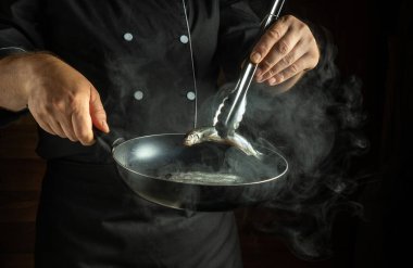 Bir şef kızartma tavasındaki taze balığı ustalıkla idare eder. Loş bir mutfağın ılık atmosferinde buhar yükselir ve lezzetli bir akşam yemeği hazırlar..