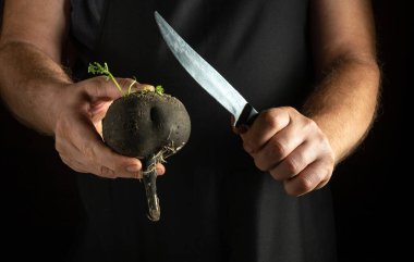 Bir insan bir elinde taze toplanmış bir siyah turpu tutarken diğer elinde keskin bir bıçak tutuyor. Ortam karanlık, hazırlık sürecine odaklanmayı arttırıyor..