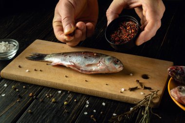 Elleri ustalıkla taze yakalanmış bir balığın üzerine baharat serpiyor. Tahta kesme tahtasına serpiştiriliyor. Yakınlarda, tuz kristalleri titreşiyor ve kırsal bir masada lezzetli bir hazırlık yapıyor..