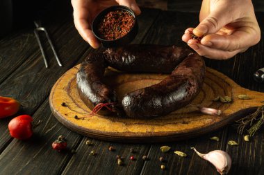 Bir şef akşam yemeğinde servis etmeden önce kanlı sosise kuru aromatik baharatlar ekler. Sosis, aromatik baharatlar ve biberlerle çevrili ahşap bir kesme tahtasının üzerinde duruyor..