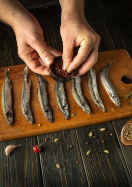 Taze balıklar ahşap bir tahtanın üzerine dizilir. Elleri aromatik baharatlar serpiştirir. Kır manzarası, yemek pişirme sanatını ön plana çıkaran lezzetli bir yemeğin otantik hazırlığını vurguluyor..