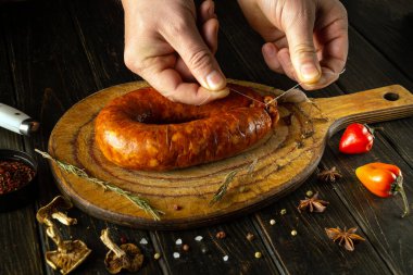 Eller ustalıkla yeni yapılmış bir sosisi ahşap bir tahtaya bağlıyor. Canlı biberler ve mis kokulu bitkiler lezzetli yaratılış, gösterişli aşçılık sanatı ve geleneksel yöntemleri çevrelemektedir..