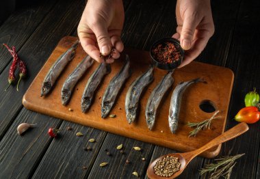 Eller, ahşap bir tahtanın üzerine serpilmiş taze balıklara ustalıkla baharat serpiyor. Lezzetli bir yemek için hazır malzeme ve lezzetlerin canlı bir karışımını sergiliyor..
