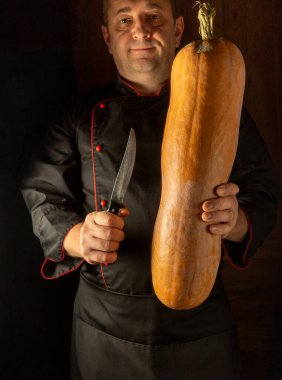 A skilled chef in a black uniform shows off a huge butternut squash, preparing to transform it into a delicious dish with a knife. Warm lighting adds to the cozy atmosphere. clipart