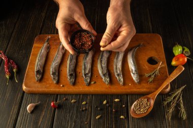 Hands skillfully sprinkle aromatic spices over a line of fresh fish on a wooden cutting board. Colorful chilies and herbs surround the setup, showcasing a vibrant culinary preparation. clipart