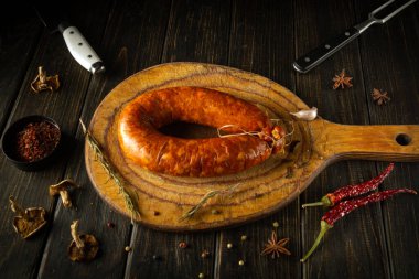 Çok güzel işlenmiş bir sosis, canlı baharatlar ve bitkilerle çevrili, kırsal kesim tahtasının üzerinde duruyor. Sıcak tonlar bu rahat mutfak ortamında davetkar bir atmosfer yaratıyor..