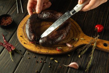 Eller, baharatlar ve sarımsaklarla çevrili, ahşap bir tahtanın üzerinde el yapımı, koyu bir sosisi dikkatlice dilimleyip sıcak bir mutfak atmosferinde mutfak işçiliğinin özünü yakalıyor..