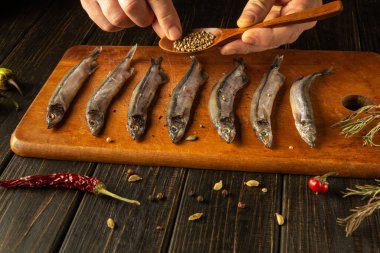 Eller ahşap kesme tahtasına dizilmiş taze hazırlanmış balıkların üzerine aromatik baharatlar serperler. Köy ortamında bitkiler ve biberler var. Mutfak etkinliğine sıcaklık katıyor..