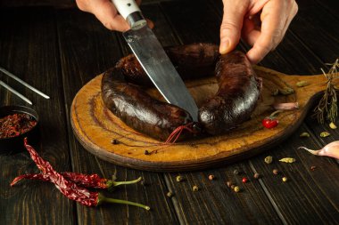 Yetenekli bir şef ahşap kesme tahtasındaki lezzetli bir sosisi dilimlemeye hazırlanır. Taze otlar ve baharatlar yemeğin etrafını sarar. Mutfak ortamındaki mutfak deneyimini geliştirir..