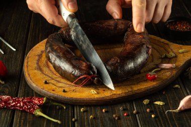 Eller, baharatlar ve sarımsaklarla çevrili, ahşap bir tahtanın üzerinde el yapımı, koyu bir sosisi dikkatlice dilimleyip sıcak bir mutfak atmosferinde mutfak işçiliğinin özünü yakalıyor..