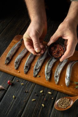 Yetenekli eller, ahşap kesme tahtasındaki bir sıra taze tutulan balığın üzerine nazikçe baharat serpiyor. Sıcak, kırsal bir ortam aşçılık hazırlığını güçlendirir, yemek pişirme sanatını ön plana çıkarır..