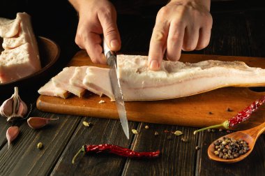 Elleri, canlı baharatlar ve sarımsak ile çevrili ahşap bir tahtanın üzerindeki leziz domuz göbeğini ustalıkla kesiyor. Mutfağın sıcak havası bu mutfak anını güzelleştiriyor..