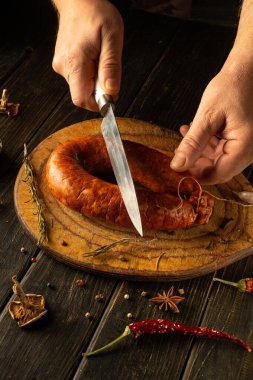 Yetenekli eller, ahşap bir tahtanın üzerinde güzel tütsülenmiş bir sosisi dilimler. Baharatlar ve otlar lezzetin etrafını sarar. Geleneksel bir hazırlık metodu sergilerler..