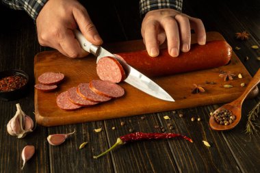 Eller ahşap bir kesme tahtasıyla çeşitli sosisleri ustalıkla dilimler. Baharatlar, sarımsak ve canlı biberler yemek pişirmek için sıcak ve davetkar bir ortam yaratıyor..