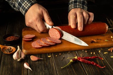 Elleri, ahşap bir tahtanın üzerinde, renkli baharatlar, sarımsak ve kırmızı biberlerle çevrili zengin bir sosisi ustalıkla dilimliyor. Mutfak ortamında mutfaktaki yaratıcılığın özünü yakalıyor..