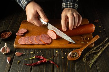 Eller ahşap bir tahtanın üzerinde, taze sarımsak, otlar ve canlı baharatlarla çevrili zengin bir sosisi ustalıkla dilimler, rahat bir mutfak ortamında aşçılık yaratıcılığının özünü canlandırır..