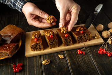 Bir çift el, ceviz ve kırmızı böğürtlenleri taze kesilmiş cevizli kekin üzerine dikkatlice yerleştirir. Kurulumda, sıcak ve davetkar bir atmosferi çağrıştıran kırsal ahşap bir masa yer alıyor..