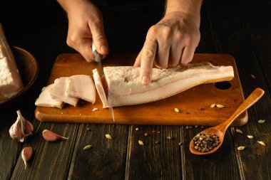 Eller, ahşap bir kesme tahtasındaki taze domuz göbeğini ustalıkla keser. Sarımsak karanfilleri ve baharatlar yakınlarda sanatsal olarak düzenlenir ve kırsal mutfağın mutfak atmosferine eklenir..