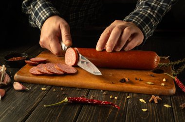 Yetenekli eller leziz bir sosisi dikkatlice dilimlerken aromatik baharatlar ve taze sarımsaklarla çevrili. Sıcak, davetkar mutfak yaratıcılığı ve gelenek gösterileri.