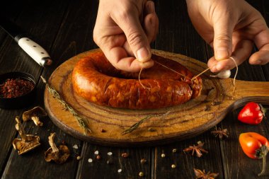 Eller, canlı otlar ve baharatlarla çevrili ahşap bir tahtanın üzerinde ev yapımı sosisi ustalıkla hazırlar. Mutfağın sıcaklığı mutfak yaratıcılığı için davetkar bir sahne hazırlıyor..