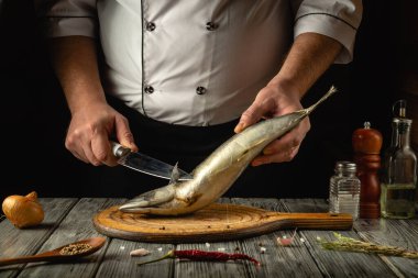 Yetenekli bir şef ahşap kesme tahtasındaki taze balığı ustalıkla fileto yapar. Soğan, baharat ve yağ gibi etrafını saran malzemeler kırsal bir mutfakta davetkar bir atmosfer yaratır..