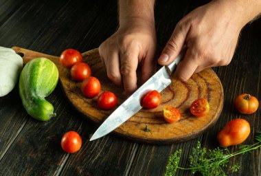 Eller, salatalık ve bitki gibi taze sebzelerle çevrili yuvarlak bir tahta üzerinde ustalıkla olgun domatesleri doğrarlar. Sıcak öğleden sonra ışığı ürünlerin canlı renklerini vurgular..