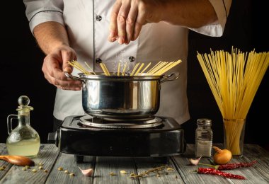 Yetenekli bir şef, soğan ve otlar gibi taze malzemelerle çevrili kaynayan bir tencereye makarna serpiyor. Yemek pişirme seansı sırasında aşçılık sanatını sergiliyor..