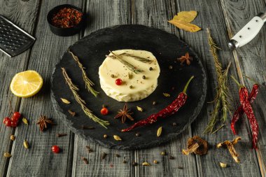 Gri bir mutfak masasında, üzerinde otlar ve baharatlar olan yuvarlak bir peynir sergileniyor..