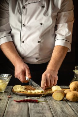 Eller patatesleri ahşap bir yüzeyde ustalıkla doğruyor, yakınlardaki alet ve malzemelerle. Kırsal mutfağın sıcak atmosferi yemek pişirme tutkusunu vurgulayarak odaklanmış aşçılık sürecini geliştirir..