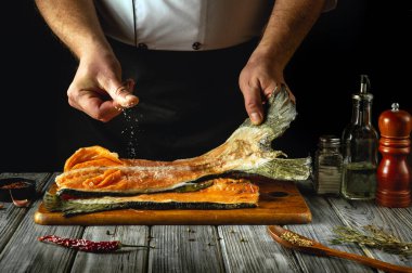 Becerikli bir şef, taze somon balığı ahşap bir kesme tahtasının üzerinde dinlenirken, üzerine tuz ve otlar serpiştirir. Zeytinyağının ve baharatların ılık parıltısı atmosferi güçlendirir..