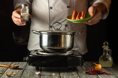 Becerikli bir şef sıcak bir mutfakta duruyor, elinde bir tabak sosis tutarken tencereye tuz ekliyor. Buhar yükselir, aşçılık sanatıyla dolu davetkar bir atmosfer yaratır..