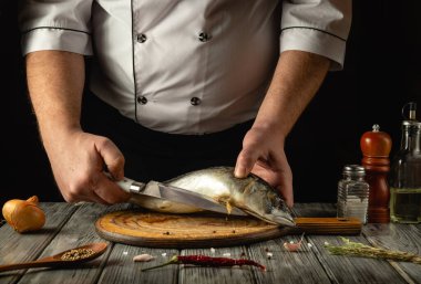 Kaba bir mutfakta, yetenekli bir şef, ahşap bir kesim tahtasıyla taze balığı ustalıkla dilimliyor, hassasiyet ve sanatsallık sergiliyor. Malzemeler ustalıkla düzenlenmiş, mutfak atmosferini güçlendiriyor..