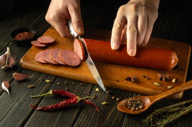 Sıcak, davetkar bir mutfakta, elleri ahşap bir kesme tahtasındaki sosis kütüğünü ustalıkla kesiyor. Etrafını saran baharatlar ve sarımsak canlı bir mutfak atmosferi yaratır. Yemek severler için mükemmel..