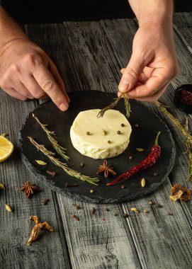 Eller dikkatlice kekik ve canlı baharatları sade ahşap bir masada kremalı peynir tekerinin etrafına dizer. Taze malzemeler mutfak sunumu sanatını geliştirir.