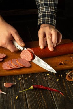 Eller, canlı baharatlarla çevrili ahşap bir kesme tahtasının üzerinde lezzetli bir sosisi ustalıkla dilimler. Sıcak ortam, aşçılık zanaatkarlığını ve iyi yemek hazırlama tutkusunu ön plana çıkarıyor..