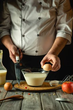 Sıcak, kırsal bir mutfakta, bir şef bir kase süte yumurta kırarak ustalıkla bir karışım hazırlar. Ahşap yüzey, mutfak atmosferini geliştiren malzemeler sergiliyor..