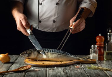 Bir şef, ahşap bir tahtanın üzerinde taze bir balığı ustalıkla fileto yaparak mutfak yeteneğini sergiliyor. Kırsal mutfak atmosferi yemek yapma sanatını ön plana çıkarıyor. Etrafında baharatlar ve çatal bıçaklar var..