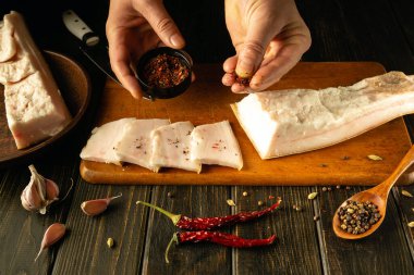 Eller dikkatlice domuz eti dilimlerinin üzerine baharat serperler, ahşap bir masada sarımsak ve kırmızı biberlerle çevrili. Sıcak ışık, mutfak yaratıcılığı için mükemmel sıcak bir atmosfer yaratır..