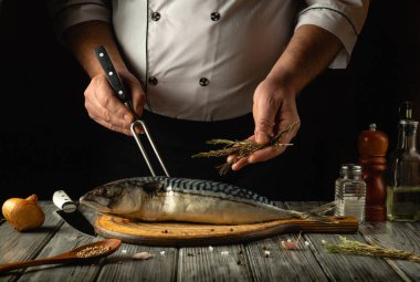 Bir şef, ahşap kesme tahtasına bütün bir balığı ustalıkla hazırlıyor, taze otlar ve baharatlar ekliyor. Sıcak mutfak atmosferi davetkar bir atmosfer yaratıyor. Mutfak macerası için mükemmel..