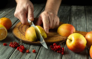 Bir çift el, narenciye ve böğürtlenlerle çevrili ahşap bir tahtanın üzerinde ustalıkla parlak elmalar keser. Sıcak renkler ve dokular mutfakta hoş, davetkar bir atmosfer yaratıyor..