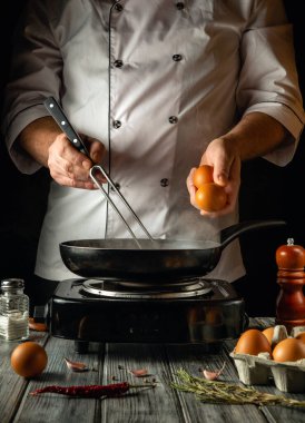 Yetenekli bir aşçı taze yumurtaları cızırdayan bir tavaya sokmaya hazırlanır. Mutfak aletleri ve malzemelerle çevrilidir. Sıcak ışıklandırma ve kırsal ortam mutfak ortamını geliştirir..