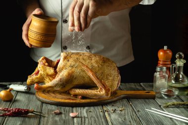 Beyaz önlüklü yetenekli bir şef, yeni hazırlanmış bir ördeğin üzerine tuz serpiyor. Malzemeler ve mutfak aletleri sıcak ve davetkar ortamda zarif bir şekilde düzenlenmiştir..