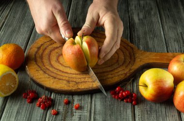 Eller renkli meyvelerle çevrili ahşap bir tahtanın üzerinde parlak bir elmayı dikkatlice dilimler. Tazelik ve aşçılık yaratıcılığıyla dolu rahat bir mutfağın imajları..