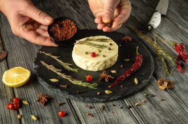 Eller, taze bitkiler, limon ve süslemelerle çevrili yuvarlak peynir çarkının üzerine ustalıkla canlı baharatlar serpiyor. Kırsal ahşap masa mutfak sanatını ve anın sıcaklığını arttırır..