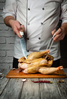 Yetenekli bir şef, taze otlar ve baharatlarla çevrili ahşap bir tahtaya çiğ tavuk keserken dikkatle odaklanır. Mutfağın sıcak parıltısı mutfak atmosferini güçlendiriyor..