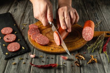 Tahta kesme tahtasında ustalıkla salam doğrayan bir çift el. Etin etrafında çeşitli baharatlar ve otlar var. Mutfak atmosferine kırsal bir dokunuş katıyor..