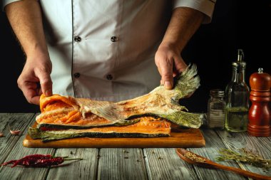 Bir şef, balığın canlı renklerini gösteren somon filetosunu ustalıkla hazırlıyor. Etrafında çeşitli çeşniler ve aletler var, rahat bir mutfakta mutfak yaratıcılığı sanatını ön plana çıkarıyorlar..