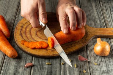 Eller ahşap bir tahtanın üzerinde parlak turuncu bir havucu ustalıkla dilimler, yanında sarımsak karanfilleri ve soğanla, sade ve ilgi çekici bir yemek atmosferi sergiler..